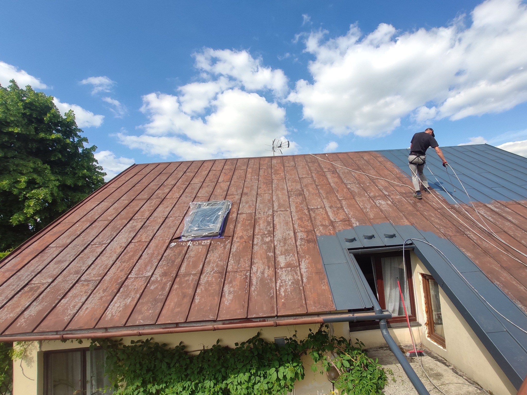 Pracownik na dachu z blachy trapezowej, częściowo pomalowanej na szaro, zabezpieczony linami, widoczne okno dachowe z folią ochronną i antena telewizyjna na tle błękitnego nieba.