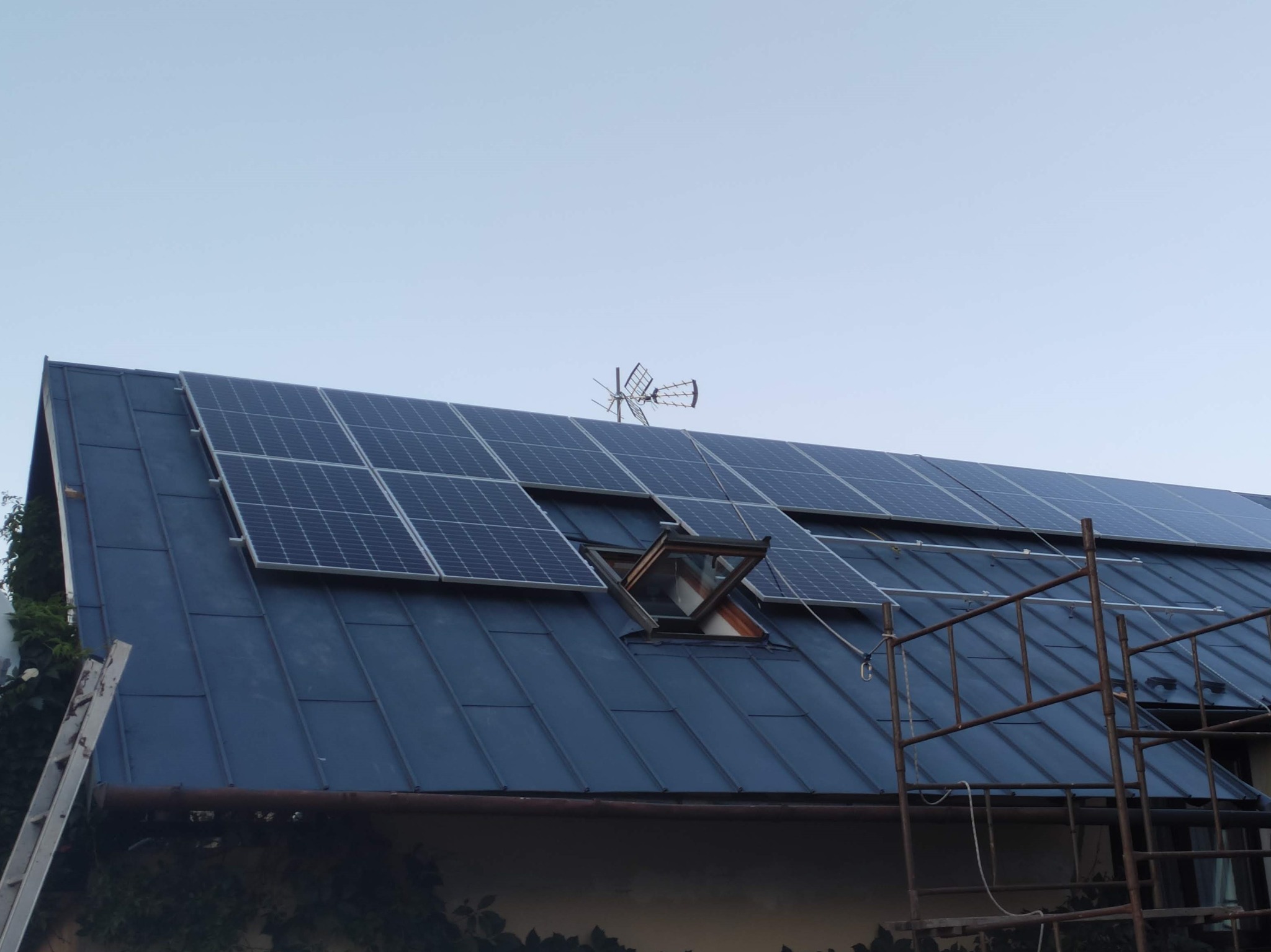 Panel fotowoltaiczny na dachu z blachy trapezowej, widoczne okno dachowe uchylone, antena telewizyjna, drabina i rusztowanie.