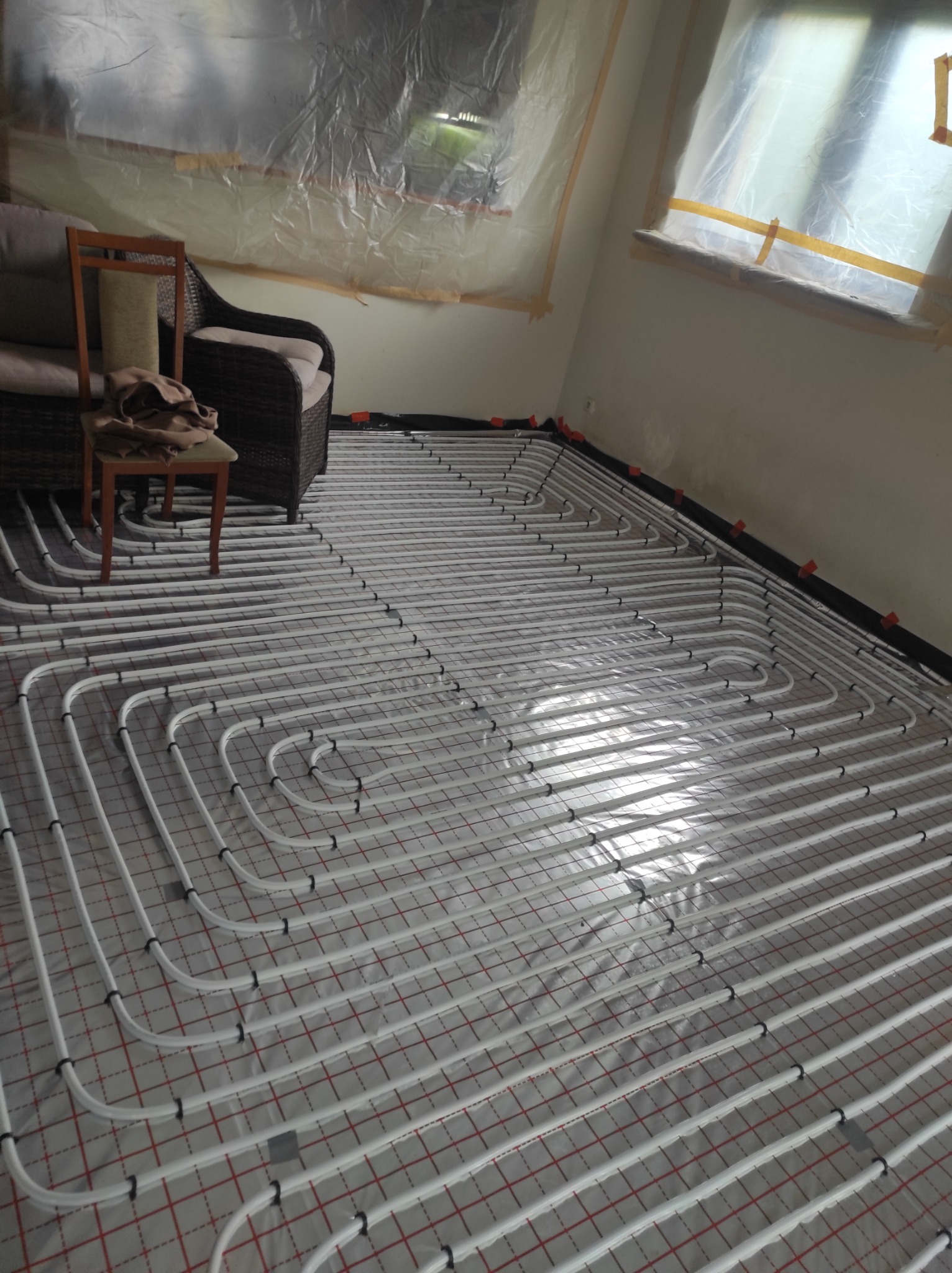 Białe rury układane w pętle na czerwono-białej siatce, tworzące system ogrzewania podłogowego w pokoju z zasłoniętymi folią oknami i meblami w rogu.