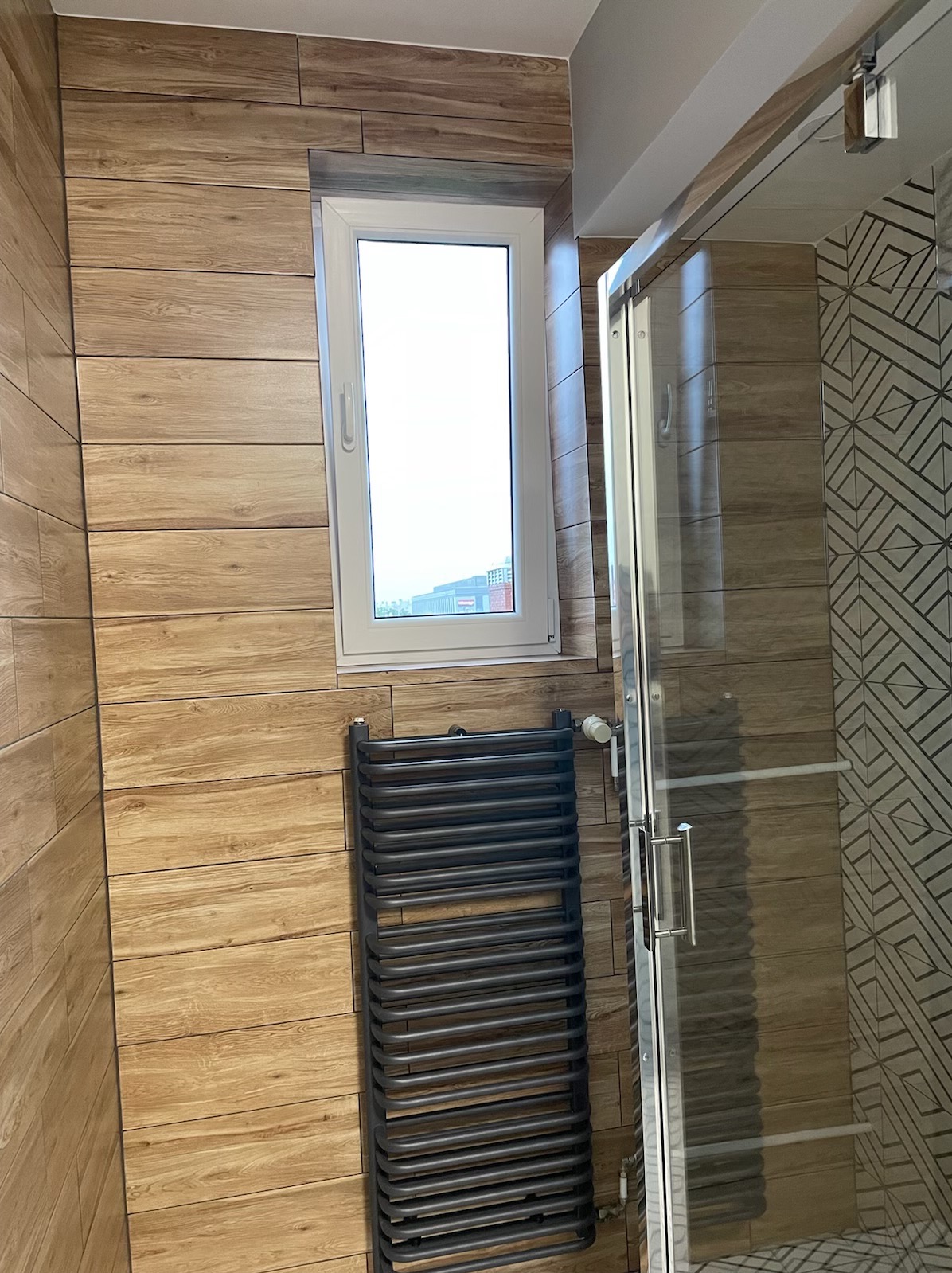 Wnętrze łazienki z kabiną prysznicową z przesuwnymi drzwiami, grzejnikiem drabinkowym w kolorze grafitowym i płytkami imitującymi drewno na ścianach oraz geometrycznymi wzorami w strefie prysznica.
