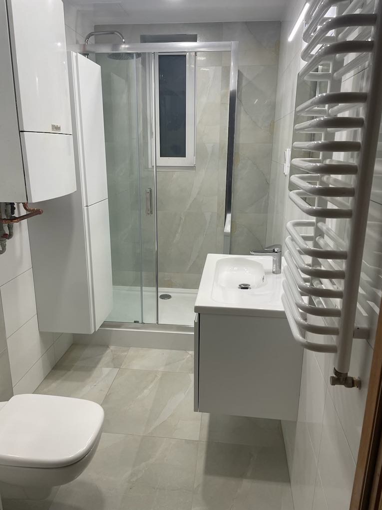 Nowoczesna łazienka w odcieniach szarości z białymi meblami wiszącymi, kabiną prysznicową z przezroczystego szkła i grzejnikiem drabinkowym, widoczny fragment toalety.