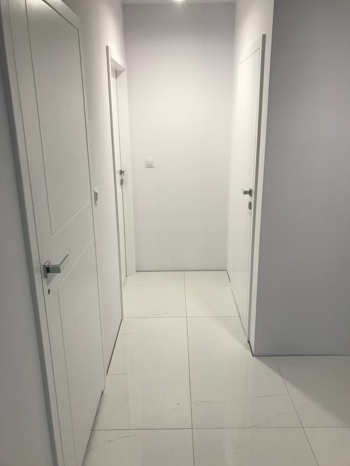Wnętrze korytarza z białymi ścianami, drzwiami i podłogą z płytek imitujących marmur, widoczne trzy pary drzwi z chromowanymi klamkami.