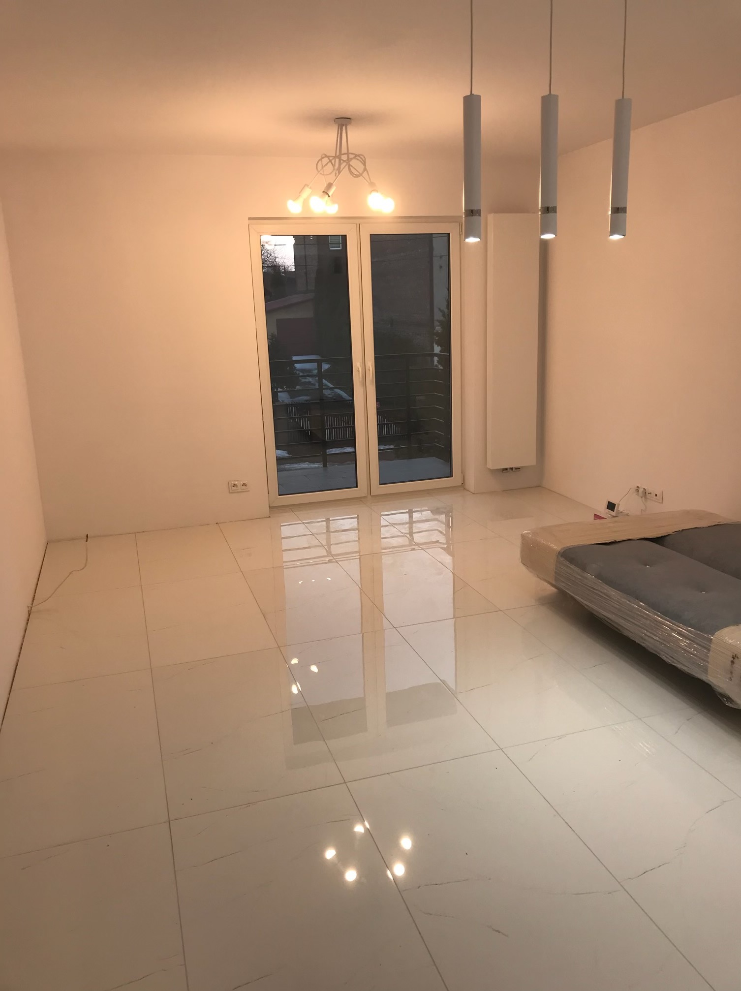 Puste, jasne wnętrze z białymi ścianami i błyszczącą podłogą z płytek, częściowo zakryte folią meble, widoczne lampy sufitowe i okno balkonowe.