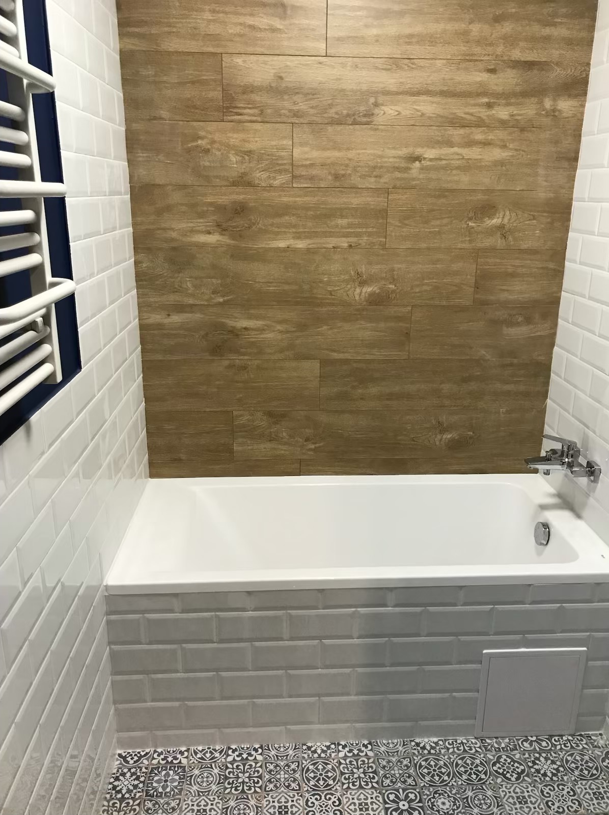 Biała wanna obudowana szarymi płytkami typu metro, z drewnopodobną okładziną ściany i dekoracyjną podłogą w łazience po remoncie.