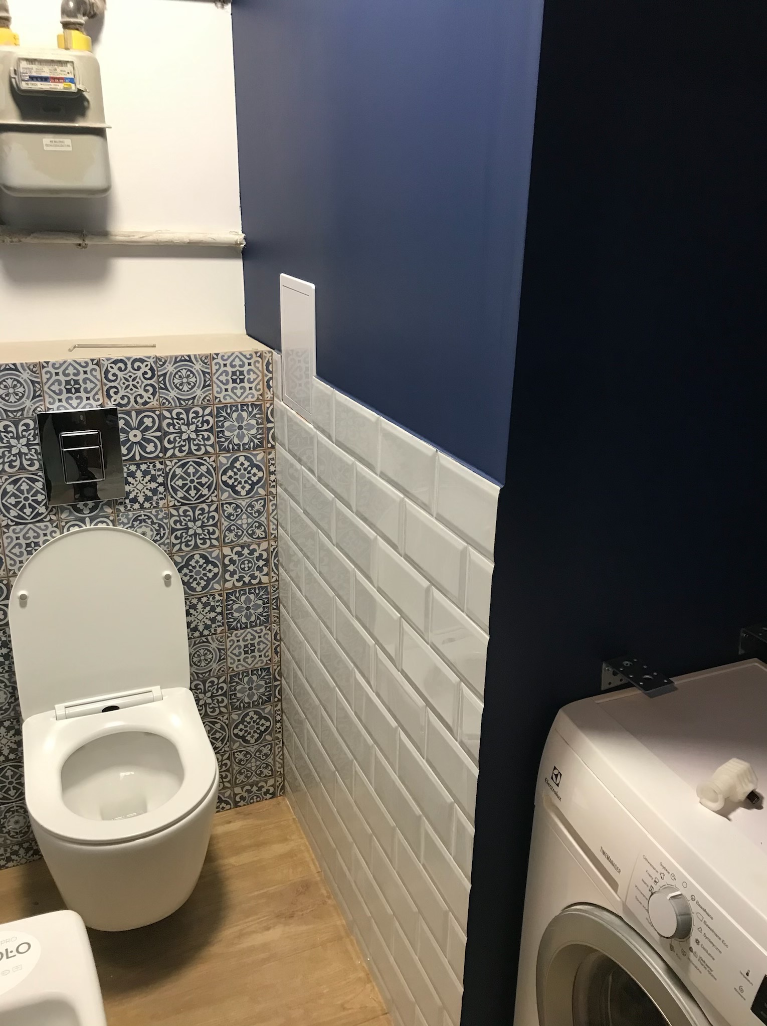 Wykończona łazienka z toaletą wiszącą, białą glazurą w stylu metro, niebieskimi płytkami dekoracyjnymi i ciemnoniebieską ścianą, widoczny licznik gazu i pralka Electrolux.