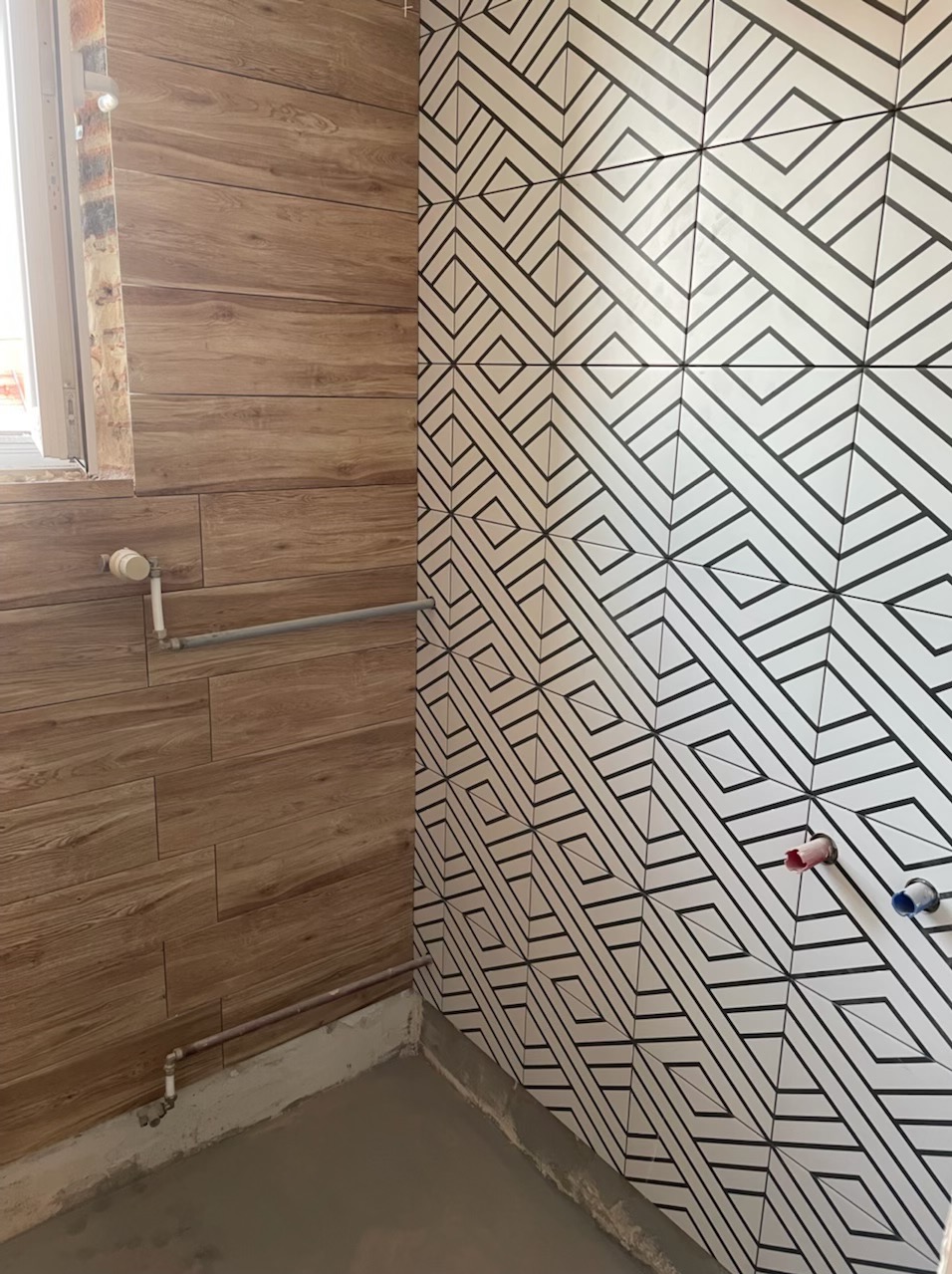 Fragment łazienki z wykończonymi ścianami: jedna z płytek imitujących drewno, druga z geometrycznym wzorem w czerni i bieli; widoczne rury instalacyjne.
