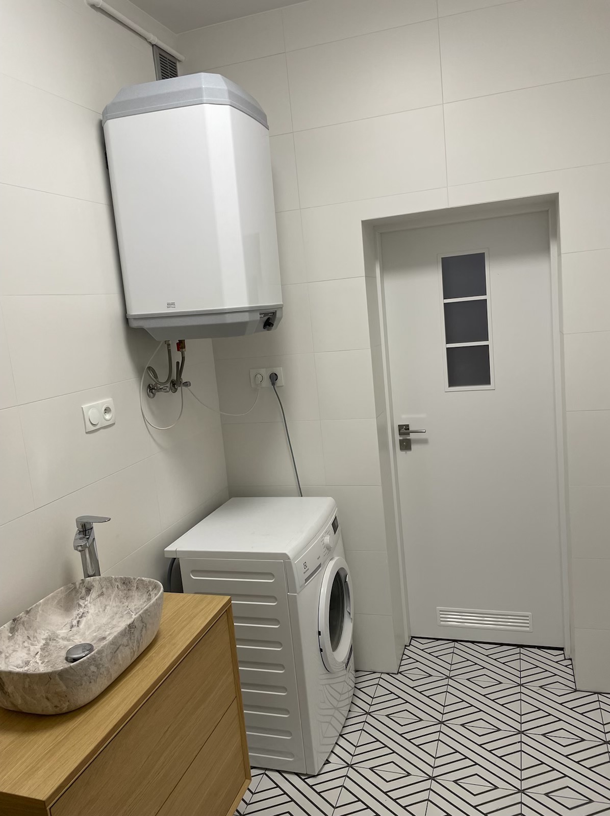 Nowoczesna łazienka z białymi płytkami, marmurową umywalką nablatową, białą pralką i wzorzystą podłogą; na ścianie zamontowany biały bojler elektryczny.