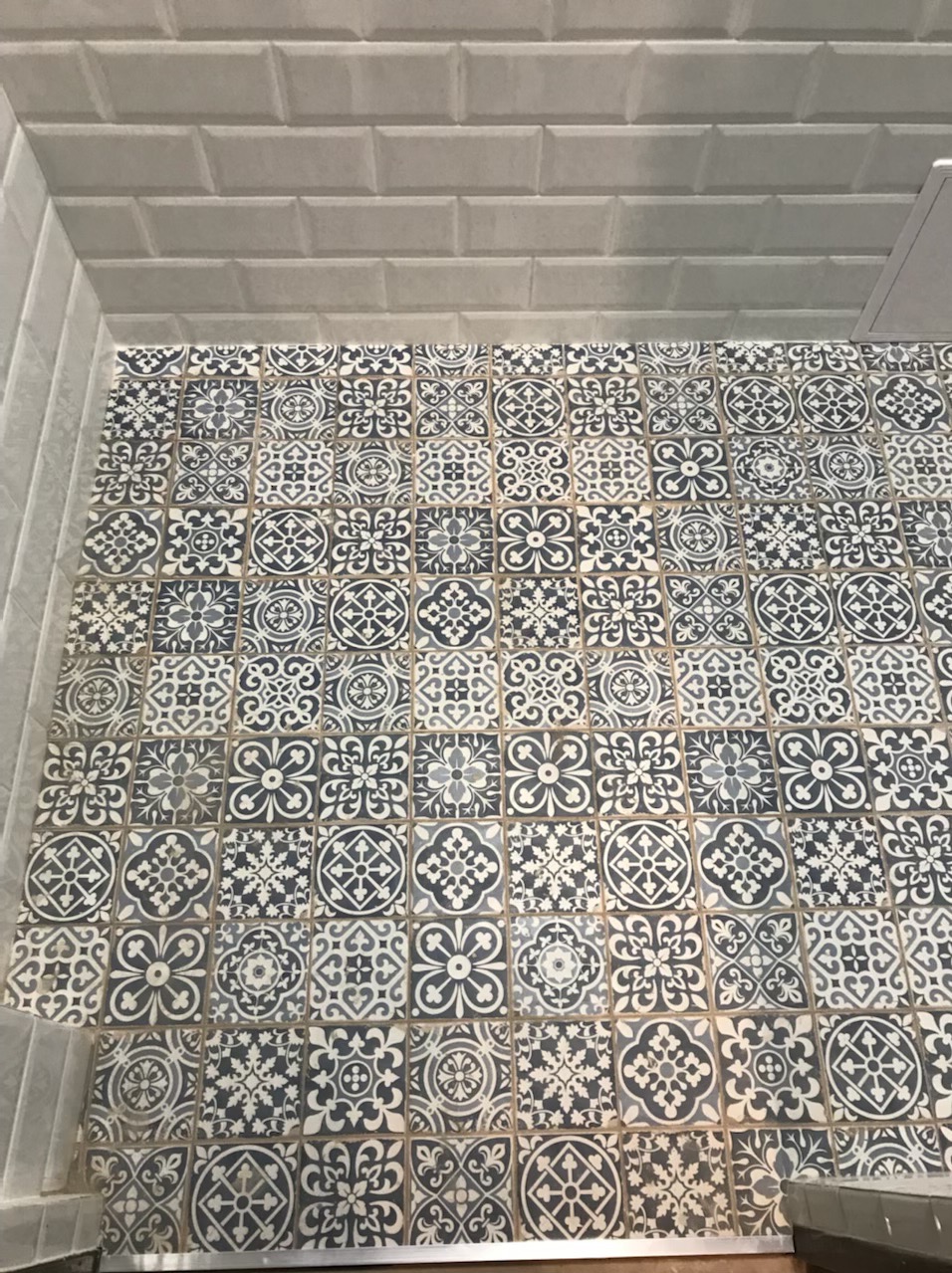 Wzorzyste, kwadratowe płytki podłogowe w odcieniach szarości i bieli, ułożone w regularny wzór, otoczone jasnoszarą, cegiełkową okładziną ścienną.