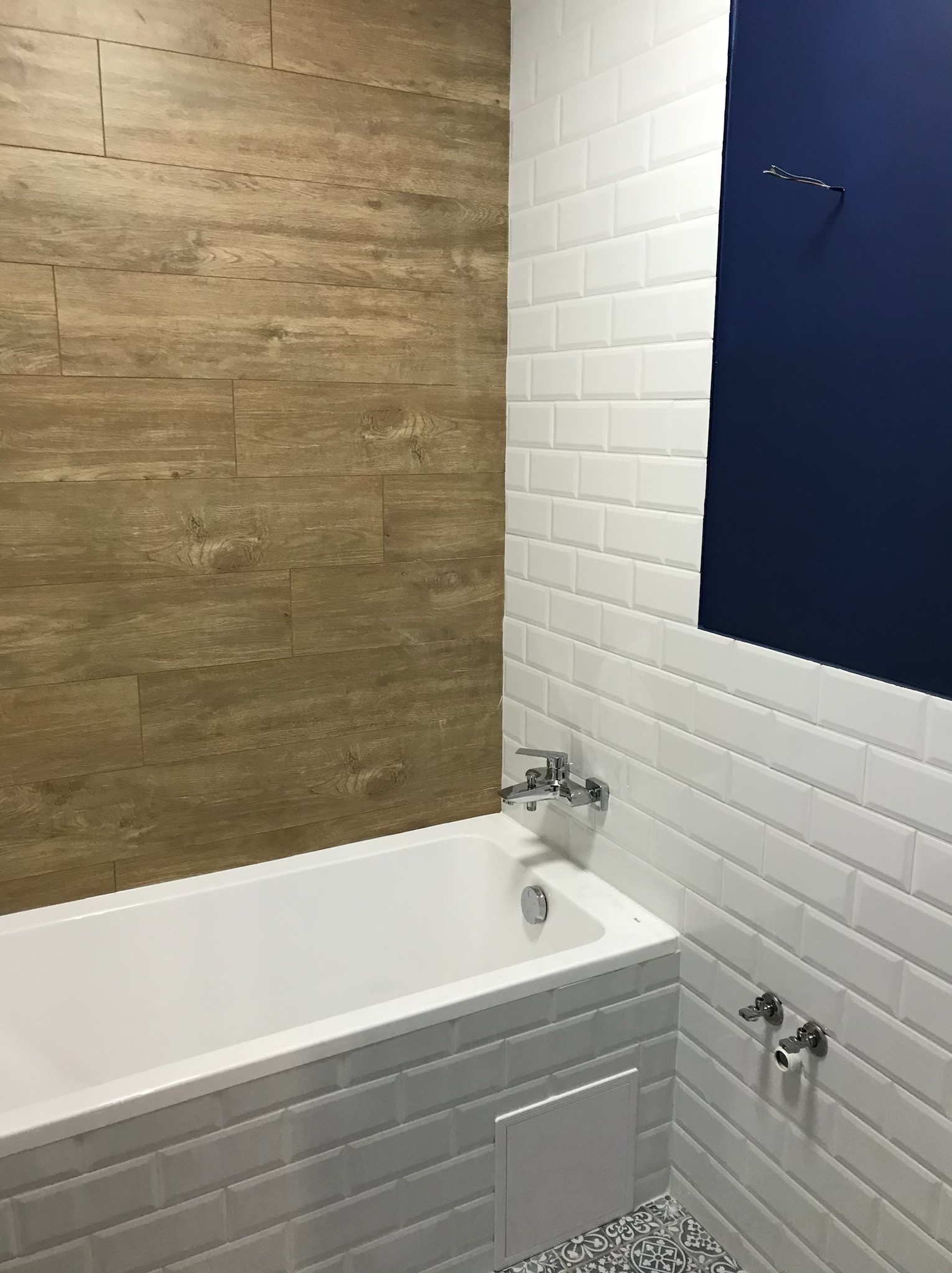 Nowoczesna łazienka z wanną obudowaną białymi kafelkami typu metro, drewnopodobną okładziną ścienną i fragmentem ściany pomalowanym na granatowo, widoczne chromowane baterie.