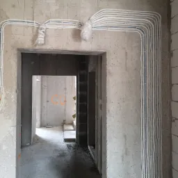 Rozprowadzone kable elektryczne w białych peszlach, częściowo zabezpieczone folią, zamontowane na surowych ścianach w nowym budynku, widoczne puszki elektryczne.