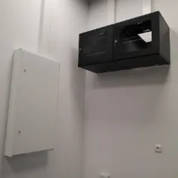 Na białej ścianie wisi czarna szafa rack z przeszklonymi drzwiami i biała skrzynka elektryczna. Widoczne gniazdka elektryczne. Minimalistyczne wnętrze.