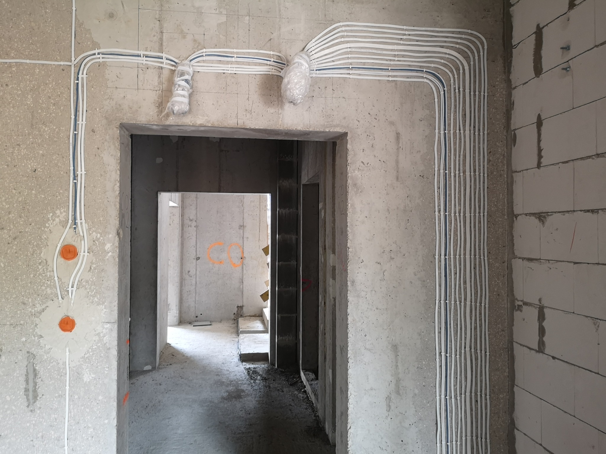 Rozprowadzone kable elektryczne w białych peszlach, częściowo zabezpieczone folią, zamontowane na surowych ścianach w nowym budynku, widoczne puszki elektryczne.