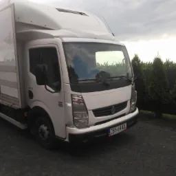 Biały samochód dostawczy Renault z zabudową kontenerową, zaparkowany na żwirowym podłożu, na tle pochmurnego nieba i zielonych tuj.