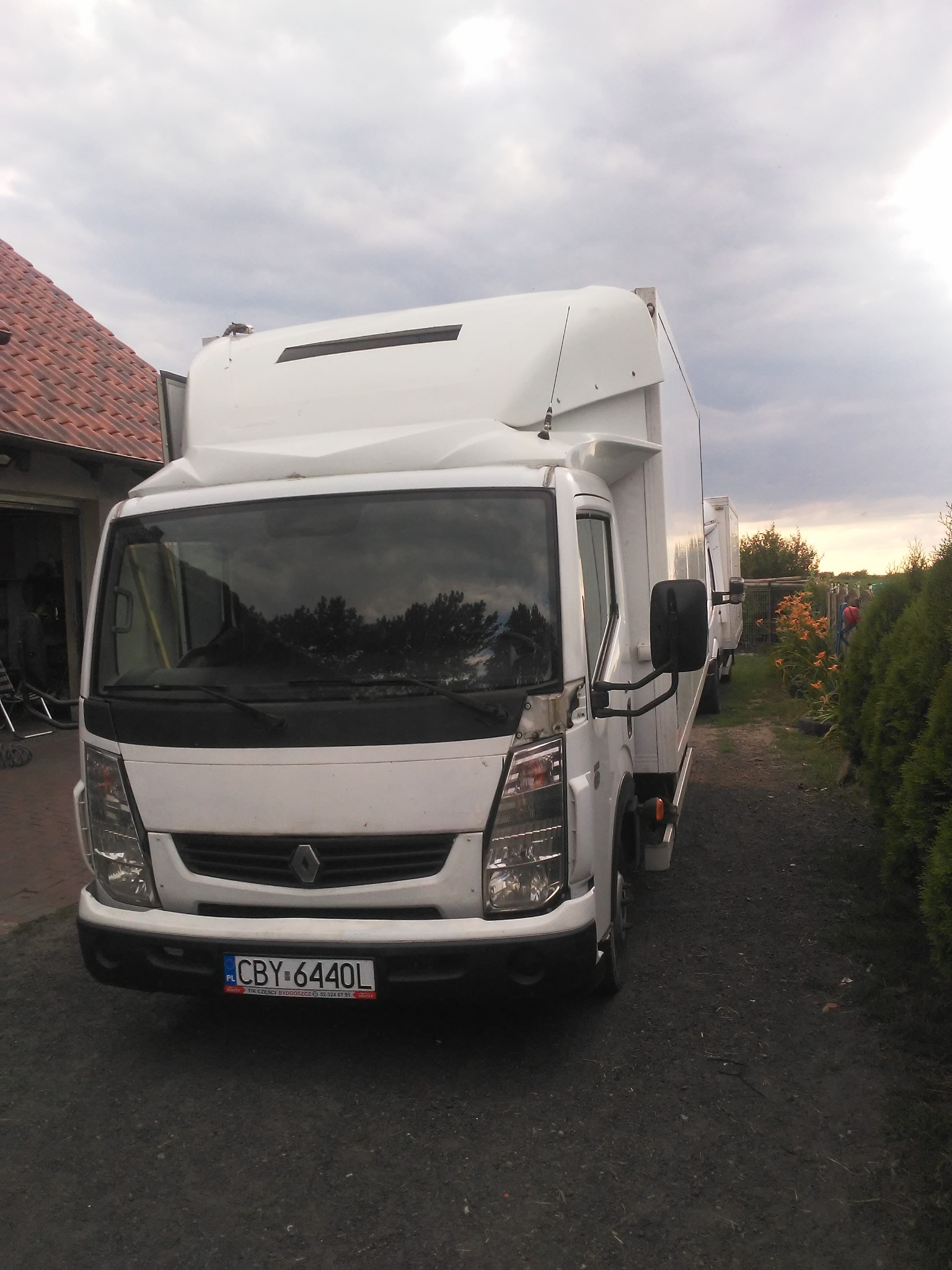 Biały samochód dostawczy Renault z zabudową kontenerową, zaparkowany na żwirowej drodze przed domem z garażem i zielonym żywopłotem w tle.