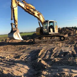 Żółta koparka Liebherr 924 podczas prac ziemnych na polu, widoczne ślady gąsienic na ziemi i usypany kopiec ziemi w tle, błękitne niebo.