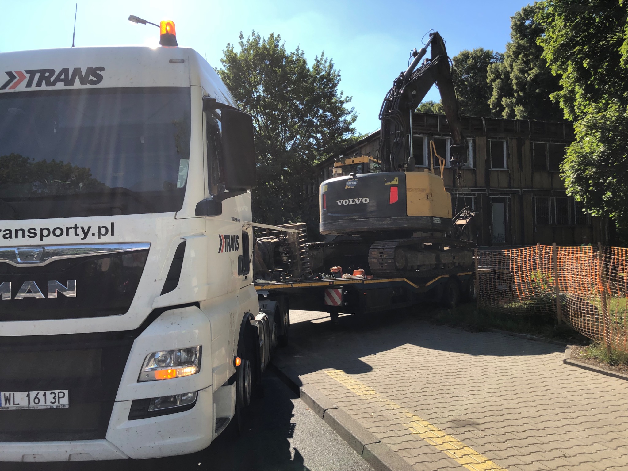 Ciężarówka transportowa z logo TRANS przewozi na lawecie żółto-czarną koparkę Volvo na tle rozbieranego budynku z pomarańczową siatką zabezpieczającą.