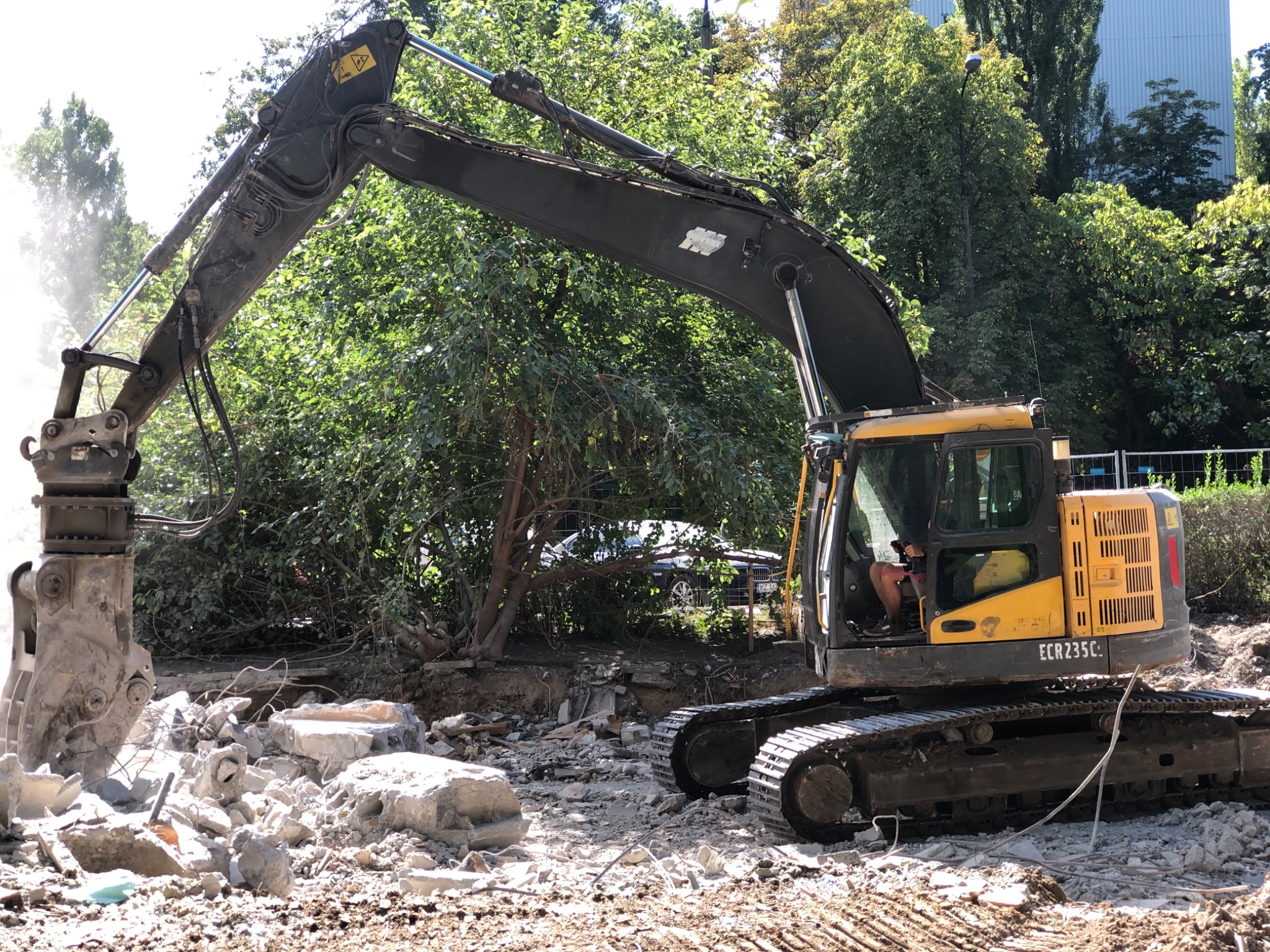 Żółto-czarna koparka Volvo ECR235C z hydraulicznym młotem kruszącym betonowe elementy na placu budowy, z zielenią w tle i fragmentem budynku.