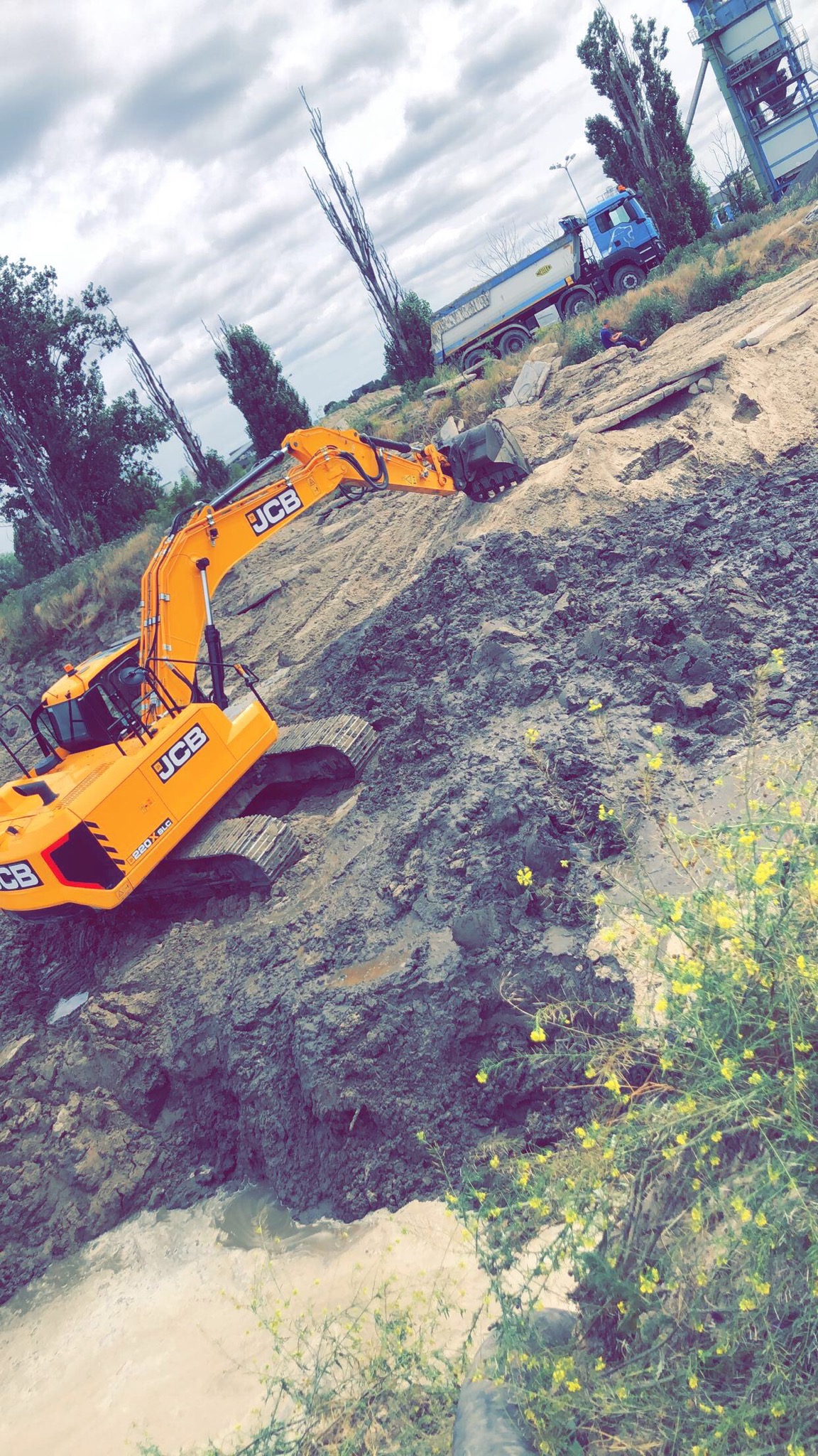 Żółta koparka JCB na nierównym, błotnistym terenie budowy, w tle wywrotka i elementy infrastruktury przemysłowej, pochmurne niebo.