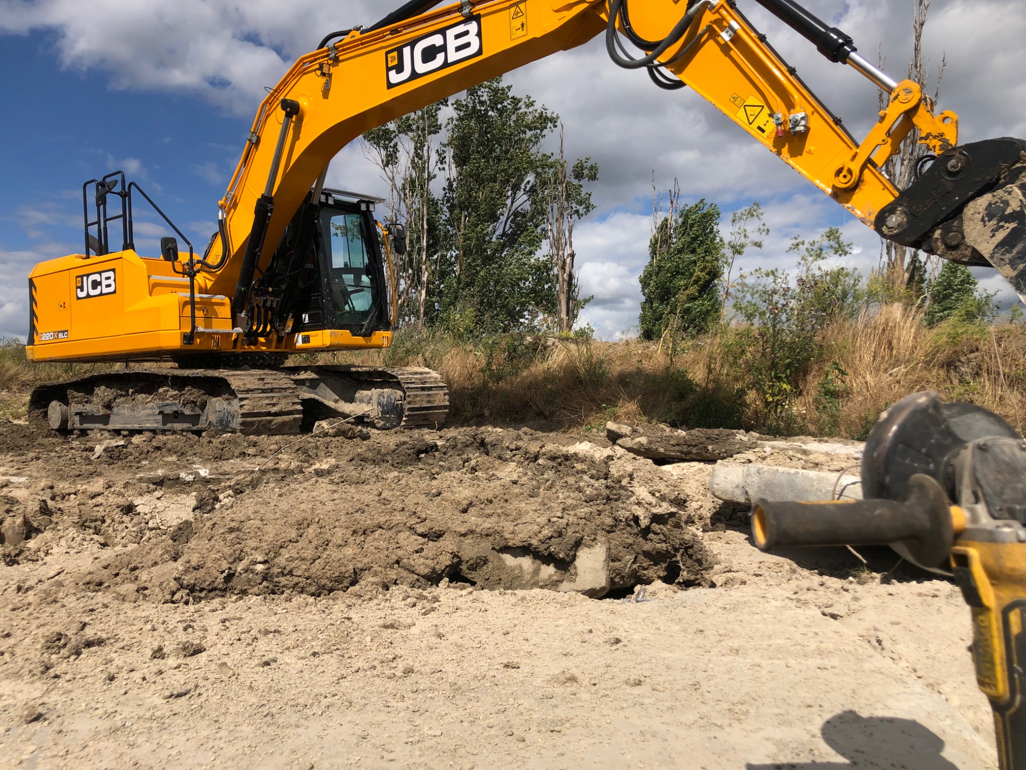 Żółta koparka JCB podczas wykopu w ziemi, z fragmentem szlifierki kątowej w prawym dolnym rogu, na tle drzew i błękitnego nieba z chmurami.