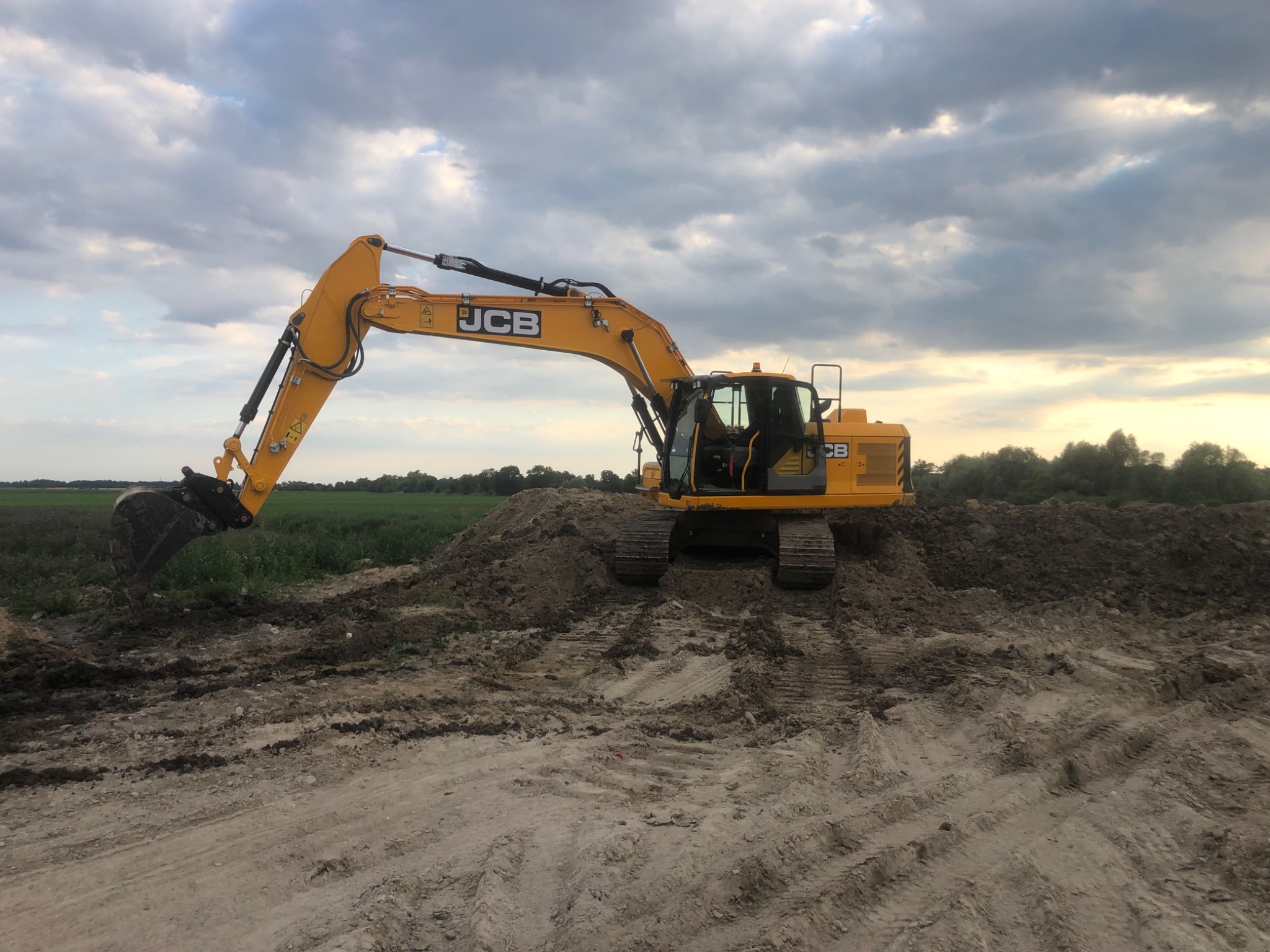 Żółta koparka JCB na nierównym terenie z widocznymi śladami opon i stertą ziemi, w tle pole i pochmurne niebo.
