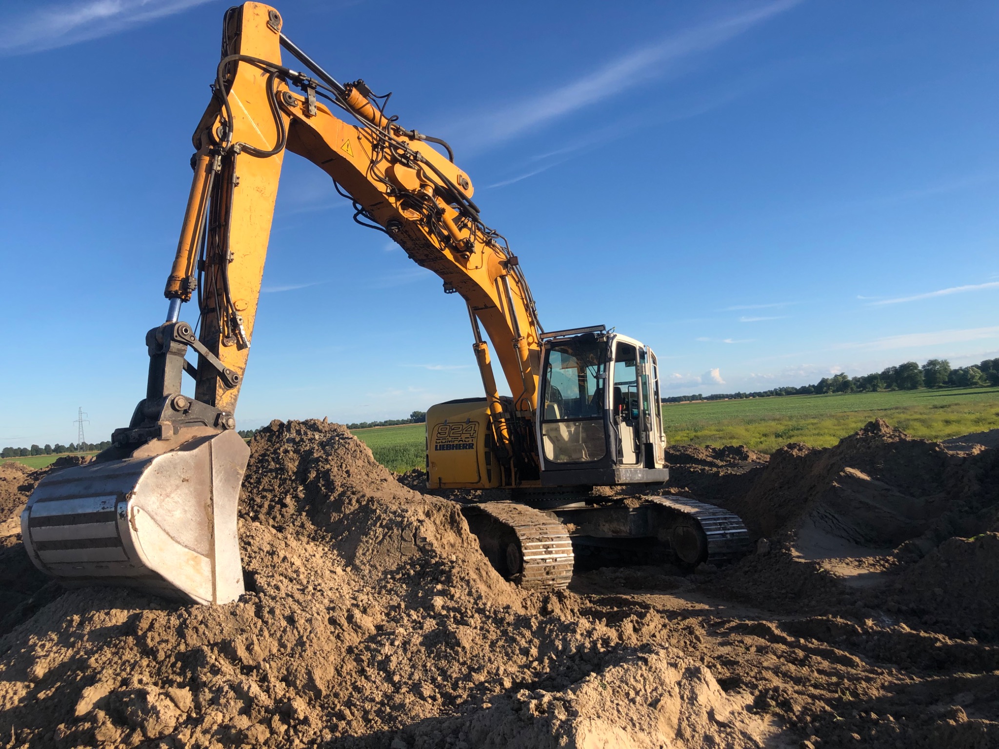 Żółta koparka Liebherr 924 Compact w trakcie prac ziemnych na polu, z widocznym wykopem i stertą ziemi, na tle błękitnego nieba i zielonych pól uprawnych w oddali.