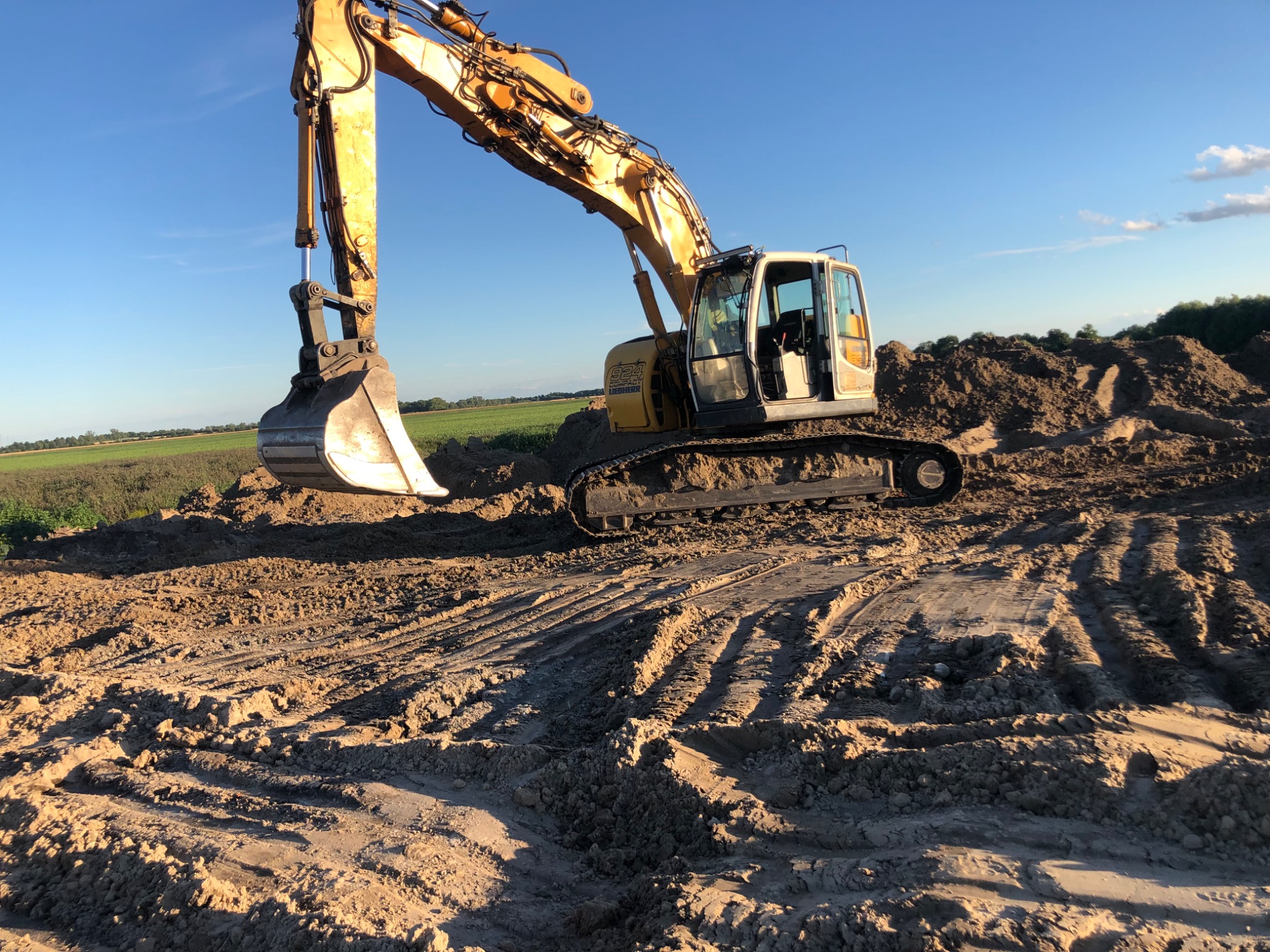 Żółta koparka Liebherr 924 podczas prac ziemnych na polu, widoczne ślady gąsienic na ziemi i usypany kopiec ziemi w tle, błękitne niebo.