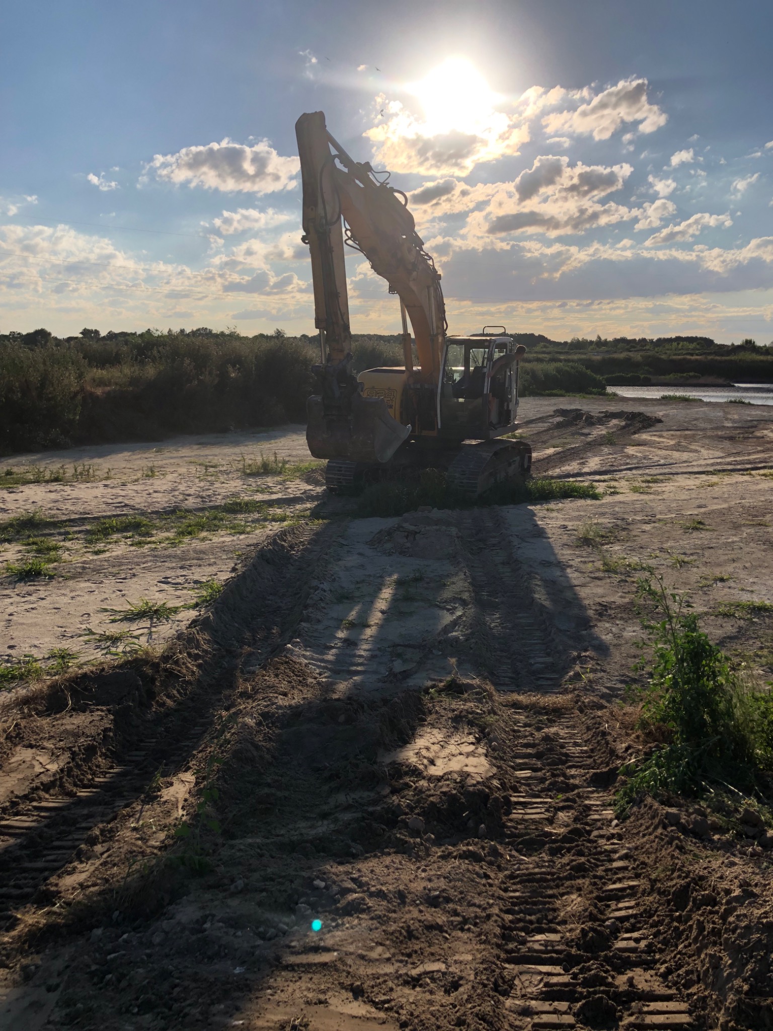 Żółta koparka gąsienicowa na piaszczystym terenie, widoczne ślady gąsienic, w tle roślinność i zachmurzone niebo z przebijającym słońcem.