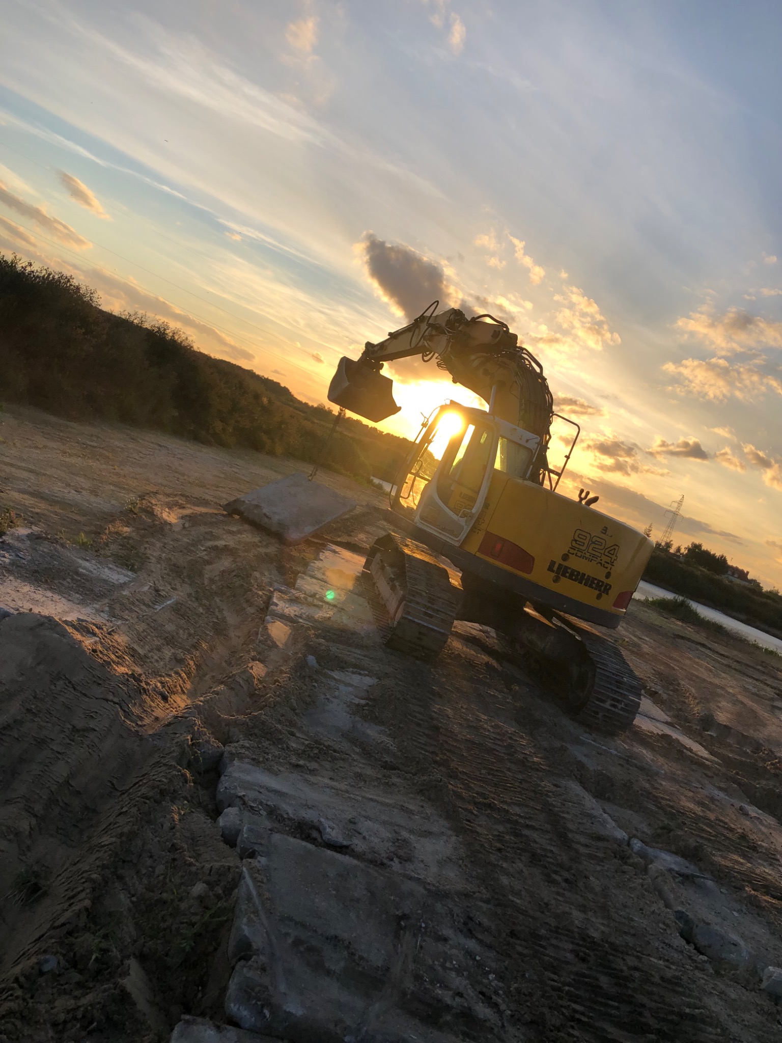 Żółta koparka Liebherr 924 Compact na placu budowy o zachodzie słońca, z widocznymi śladami gąsienic na ziemi i elementami betonowymi w tle.