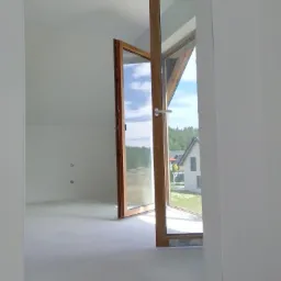 Wnętrze nowo wykończonego, jasnego pokoju z otwartym drewnianym oknem balkonowym, przez które widać dom i zieleń. Ściany i podłoga w jasnym, neutralnym kolorze, przygotowane do dalszych prac...