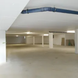 Pusta, jasna przestrzeń biurowa lub magazynowa z pomalowanymi na biało ścianami, stropem i kolumnami. Widoczne rury pod sufitem i płyty opierające się o ścianę w oddali.