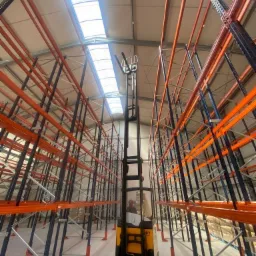 Wysoki regał magazynowy z pomarańczowymi belkami i czarnymi słupami, oświetlony przez świetliki w dachu, z widocznym wózkiem widłowym między regałami.