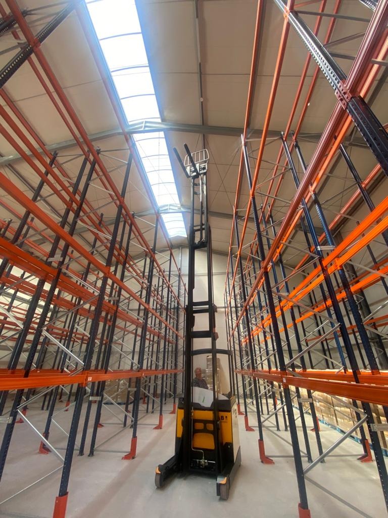Wysoki regał magazynowy z pomarańczowymi belkami i czarnymi słupami, oświetlony przez świetliki w dachu, z widocznym wózkiem widłowym między regałami.