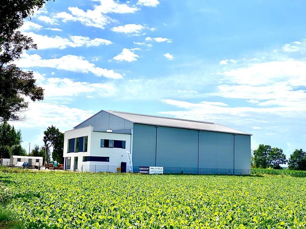 Nowoczesna hala magazynowa z biurową dobudówką, otoczona polem uprawnym w słoneczny dzień.