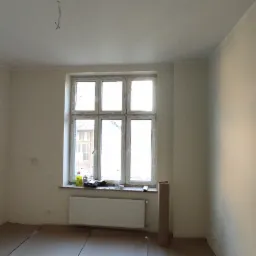 Pokój w trakcie prac przygotowawczych: ściany po gruntowaniu, zabezpieczona podłoga, widoczne kable pod sufitem i grzejnik pod oknem.