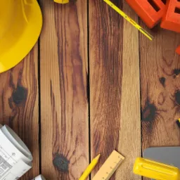 Plan architektoniczny, kask ochronny, poziomica, kielnia, cegły i inne narzędzia budowlane ułożone na drewnianym tle.