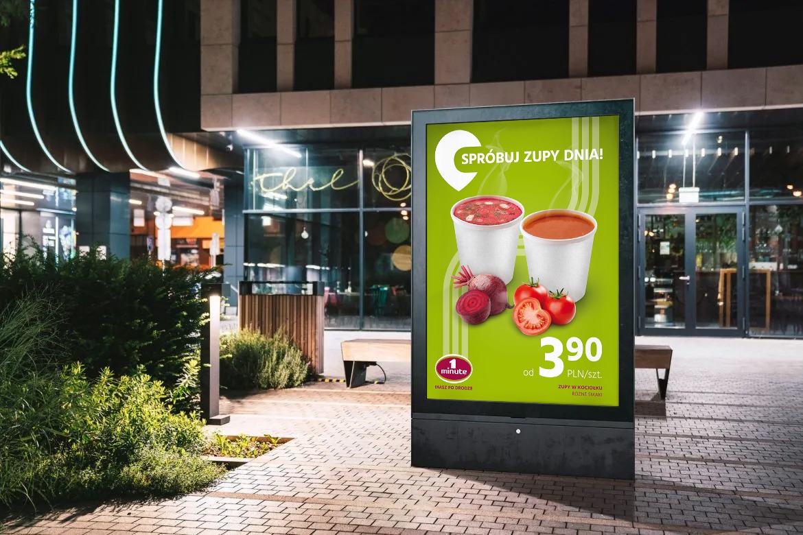 Citylight prezentujący ofertę zup na wynos w kubkach, z burakami i pomidorami, ustawiony na chodniku przed budynkiem z neonem.
