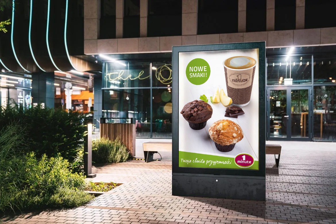 Citylight z reklamą muffinek i kawy '1 minute' na tle miejskiego budynku z neonami i kostką brukową.