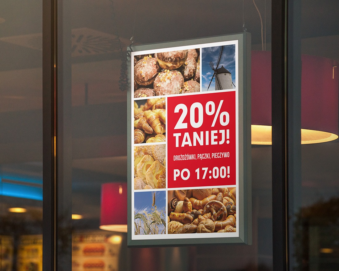 Plakat reklamowy z kolażem zdjęć pieczywa i napisem '20% taniej!' wiszący w witrynie piekarni, widoczny z ulicy.