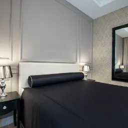 Elegancka sypialnia z panelami ściennymi w odcieniach szarości, ozdobnym lustrem w czarnej ramie i czarną pościelą z dekoracyjnym wałkiem, oświetlona lampkami nocnymi.