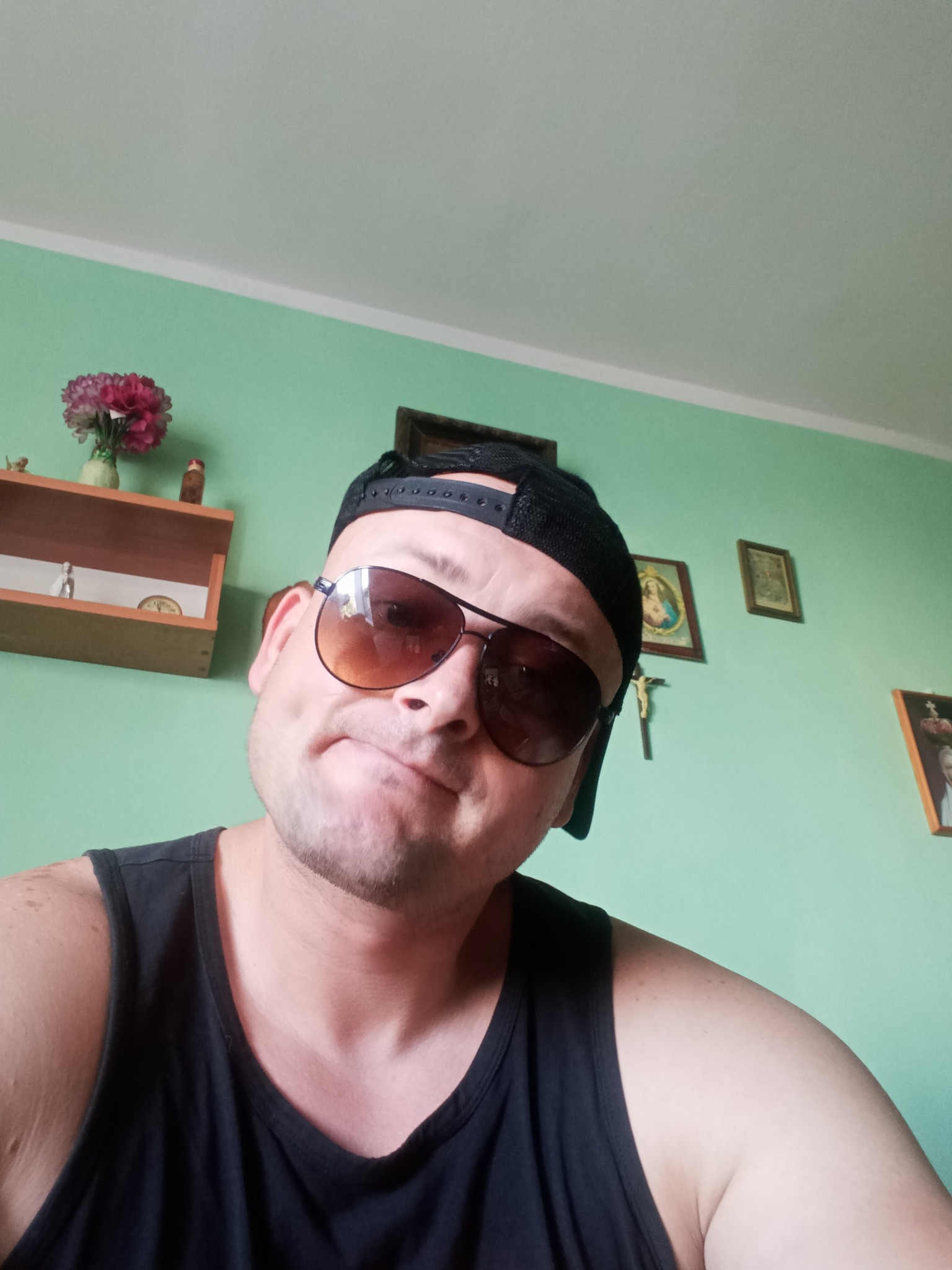 Mężczyzna w czarnej koszulce na ramiączkach i czapce z daszkiem, pozujący w okularach przeciwsłonecznych na tle miętowej ściany z półką, kwiatami i obrazkami.