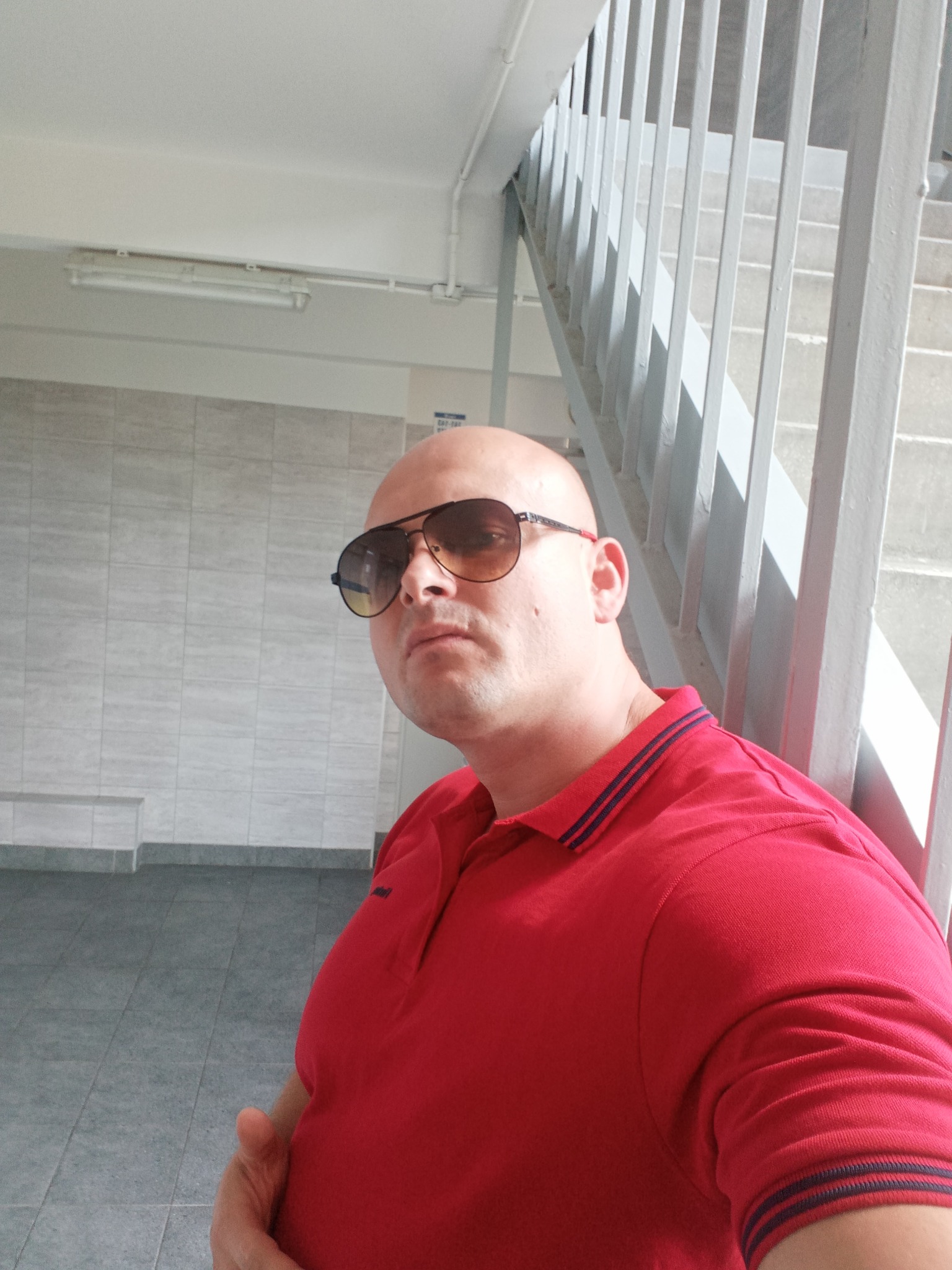 Mężczyzna w czerwonej koszulce polo i okularach przeciwsłonecznych stoi na klatce schodowej z szarą balustradą.
