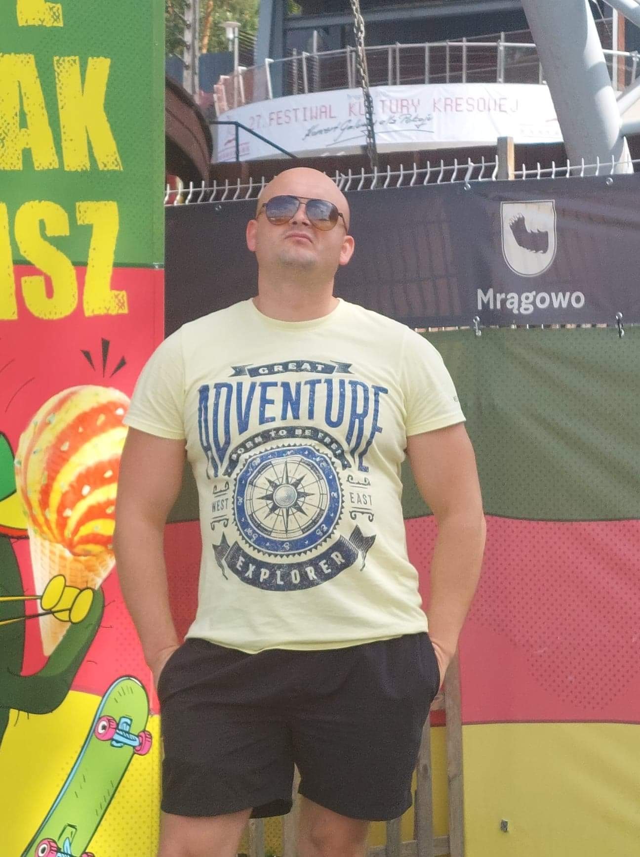 Mężczyzna w okularach przeciwsłonecznych i koszulce z kompasem na tle kolorowych banerów z napisem Mrągowo.
