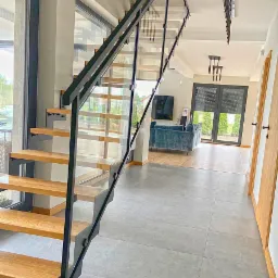 Nowoczesne schody dwubiegowe ze spocznikiem we Wrocławiu, z drewnianymi stopniami i czarną, metalową konstrukcją oraz szklanymi panelami, widoczne z dołu, z salonem w tle.