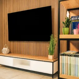 Nowoczesny salon z drewnianą ścianą z lamelami, podwieszonym telewizorem, białą szafką RTV z drewnianym blatem oraz regałem z książkami i dekoracjami roślinnymi.