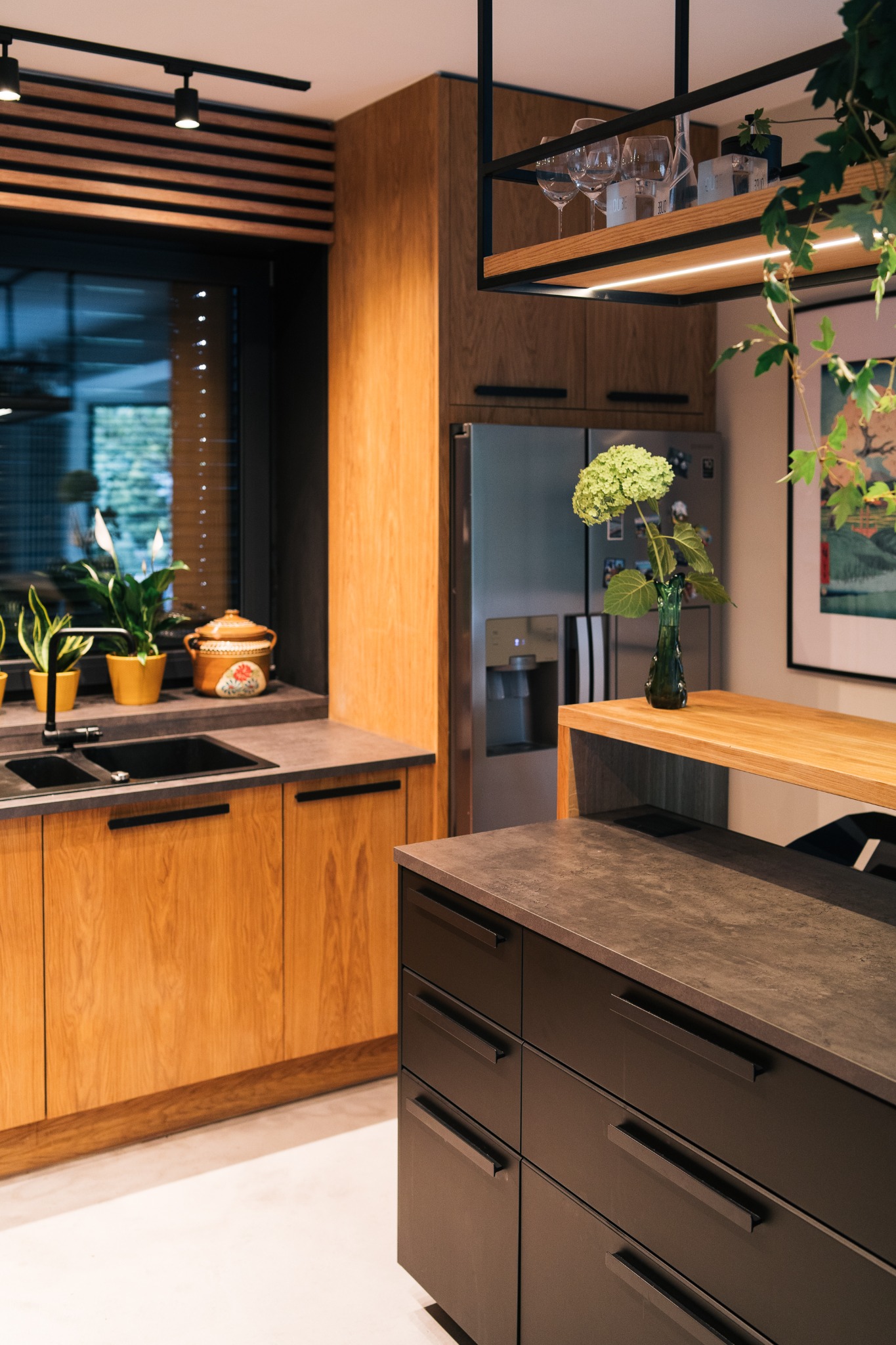 Nowoczesna zabudowa kuchenna z drewnianymi frontami, ciemnym blatem i czarnymi uchwytami, uzupełniona o rośliny doniczkowe na parapecie i kwiaty w wazonie na drewnianym blacie, oświetlona...