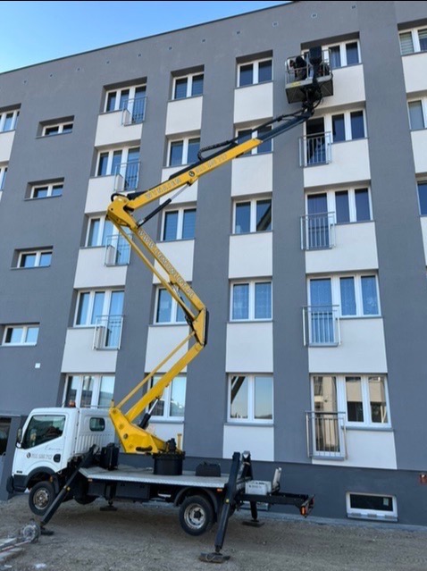 Żółty podnośnik koszowy na tle szarego budynku wielorodzinnego z białymi balkonami; pracownik w koszu podnośnika wykonuje prace na wysokości przy elewacji.