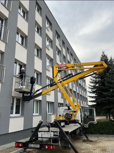 Pracownik w kasku na podnośniku koszowym maluje elewację budynku wielorodzinnego w odcieniach szarości i bieli. Widoczny fragment żółtego ramienia podnośnika z napisem 'DA 20' i numerem telefonu.