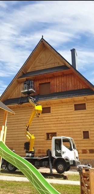 Pracownik na podnośniku koszowym maluje drewnianą elewację domu w stylu góralskim; widoczny fragment zjeżdżalni na pierwszym planie.