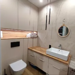 Nowoczesna łazienka z beżowymi szafkami, drewnianym blatem, białą umywalką i okrągłym lustrem w ramie z rattanu, oświetlona wiszącymi lampami tubowymi.