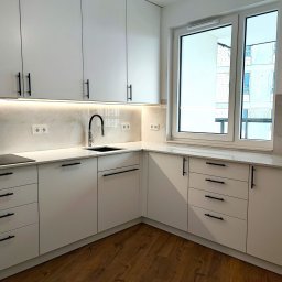 DESTO MEBLE - Nowoczesna, jasna kuchnia z białymi frontami, czarnymi uchwytami i blatem imitującym marmur, oświetlenie LED podszafkowe, widok na budynek w trakcie budowy przez okno.